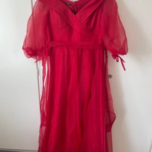 Röd off shoulder balklänning med skärp - Off shoulder balklänning med puffiga, transparenta kortärmar och omlott-detalj upptill. Klänningen har ett markerat midjeband och är tillverkad i ett lätt, flowy syntetmaterial som ger en drömmig känsla. Ärmarna går att justera, inbyggd Bh. Oanvänd. Köpt i Turkiet för 5000kr. Strl 48 men passar strl 42. Pris kan diskuteras.