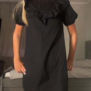 Svart kort klänning med volang - Supersöt svart kortklänning med puffärmar och stor volangkrage. Klänningen har en loose passform och är perfekt för dig som gillar en trendig och bekväm stil. Materialet känns lätt och passar bra till många tillfällen.