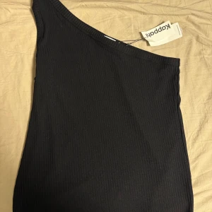 Svart oneshoulder linne från Kappahl - Snygg svart ribbad linne från Kappahl med oneshoulder-design. Linnen har en tight passform som framhäver figuren. Perfekt för dig som gillar stilrena och trendiga plagg.