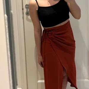 Brun/röd omlottkjol med slits - Snygg rostbrun omlottkjol med knytning och dekorativ ring i midjan. Kjolen har en lång, asymmetrisk form och en slits framtill . Köpt från marknad utomlands, använd 1 gång.