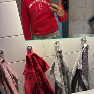 Röd Peak Performance hoodie med brodyr - Snygg röd hoodie från Peak Performance med broderad text och logga på bröstet i vitt och lila. Klassisk känguruficka framtill och huva. Perfekt för dig som gillar sportig och avslappnad stil.