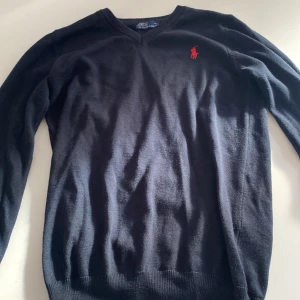 Svart stickad tröja från Ralph Lauren - Svart stickad tröja från Ralph Lauren med klassisk röd broderad logga på bröstet. V-ringad modell med långa ärmar och ribbade muddar. Perfekt för dig som gillar stilrena och tidlösa plagg med en touch av lyx.