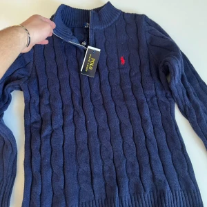 Marinblå stickad tröja Polo Ralph Lauren - Kabelstickad marinblå tröja från Polo Ralph Lauren med halv dragkedja i halsen och röd broderad logga på bröstet. Tröjan har ribbade muddar och lång ärm, perfekt för lager-på-lager under kyliga dagar.