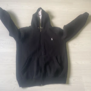 Svart hoodie från Polo Ralph Lauren - Svart hoodie från Polo Ralph Lauren storlek m  med dragkedja och klassisk vit broderad logga på bröstet. Tröjan har huva med snörning, två fickor framtill och är tillverkad i mjuk bomull. Perfekt för chill dagar eller när du vill ha en clean streetstyle. Priset är ej hugget i sten och kan förhandlas mot en snabb affär 