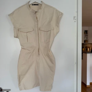Beige jeansklänning från Zara - Stilren beige kortklänning från Zara med ståkrage och två stora bröstfickor. Klänningen är ärmlös och har en rak, figurnära passform. Köpt på Sellpy!