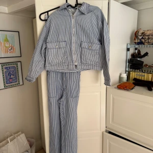 Randig jeansset i blått och vitt - Säljer en randig jeansjacka och byxdress från H&M i blått och vitt. Jackan har dragkedja framtill och är i Storlek XS, två stora fickor med lock och klassisk krage. 