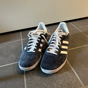 Adidas Gazelle sneakers i mörkblå mocka - Snygga Adidas Gazelle sneakers i mörkblå mocka med klassiska vita ränder och vit sula. Skorna har vit hälkappa, vita skosnören och guldfärgad Gazelle-text på sidan. 
