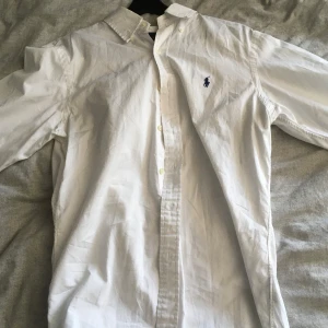 Vit skjorta från Ralph Lauren slim fit - Klassisk vit skjorta från Ralph Lauren i slim fit-modell. Skjortan har lång ärm, knappar framtill och den ikoniska blå broderade loggan på bröstet. Tillverkad i bomull och passar perfekt till en clean och stilren look.
