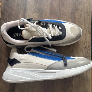 Vita sneakers med blå dragkedja från Zara - Chunky sneakers från Zara i vitt med svarta och grå detaljer samt en blå dragkedja på ovansidan. Skorna har både snörning och dragkedja, rund tå och platt sula. Materialmix av syntet och textil för en modern look.