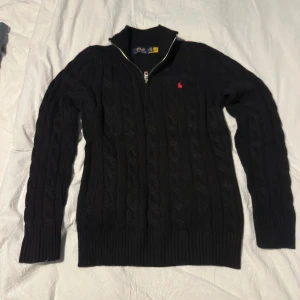 Svart kabelstickad tröja Polo Ralph Lauren - Svart kabelstickad tröja från Polo Ralph Lauren med halv dragkedja och liten röd logga på bröstet. Tröjan har ribbade muddar och hög krage, perfekt för lager-på-lager. Klassisk och stilren look som funkar till allt. Tröjan är ny och säljes pga felköp