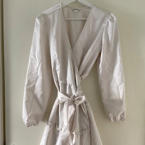  Omlottklänning från Monki - Supersöt ljus omlottklänning från Monki med långa puffärmar och knyt i midjan. Klänningen har volangdetaljer och v-ringning framtill. 