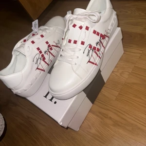 Valentino Garavani sneakers med nitar - ALDRIG ANVÄNDA! Snygga vita Valentino Garavani sneakers i skinn med röda nitar och grafiskt tryck på sidorna. Skorna har snörning, platt sula och coola detaljer som ger en unik look. Perfekta för dig som vill sticka ut med exklusiva sneakers.