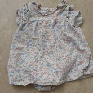Klänning - Supersöt babyklänning från H&M i ekologisk bomull. Klänningen är vit med blommigt mönster i rosa, blått och lite gult, har volangärmar och praktiska knappar i grenen. Perfekt för sommarens varma dagar!