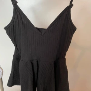 Svart ribbad linnetopp med v-ringning - Snygg svart linnetopp med v-ringning och smala axelband. Toppen är ribbad och har en lätt utsvängd nederdel som ger en cool siluett. Perfekt att styla med jeans eller kjol för en trendig look.