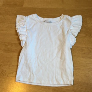 Vit ribbad topp med volangärm Zara - Supersöt vit ribbad topp från Zara med korta volangärmar. Toppen har rund halsringning och är gjord i mjuk bomull. Perfekt för dig som gillar en enkel men ändå gullig stil med extra detalj på ärmarna. Storleken är M men passar perfekt för mig som brukar ha Xs-S💕