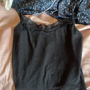 Svart ribbad linnetopp med spets - Snygg svart ribbad linnetopp med tunna axelband och spetsdetalj längs urringningen. Perfekt att styla med jeans eller kjol för en trendig look. Mjuk och skön känsla, passar till många outfits.