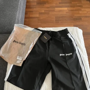 Palm Angels svarta shorts  - Svarta mjukisbyxor från Palm Angels med vita sidoränder och logga broderad på vänster ben. Byxorna har elastisk midja och sportig vibe, perfekta för en streetwear-look. Materialet är mjukt och bekvämt, och passformen är loose. Äkta såklart