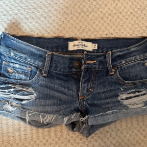 Blå jeansshorts från Abercrombie - Snygga korta blå jeansshorts från Abercrombie med slitna detaljer och fransig kant. Klassisk femficksmodell med låg midja och coola slitningar framtill och baktill. Perfekta för en avslappnad och trendig look i sommar. Midjemått 38cm 