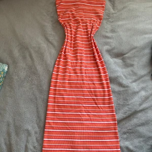 Ribbad lång tubklänning - Säljer en lång, ribbad klänning som är orange och vitt med smala horisontella ränder. Klänningen har en figurnära passform, liten slits där bak och är ärmlös. Perfekt för dig som gillar färg och vill sticka ut lite extra. Aldrig använd 