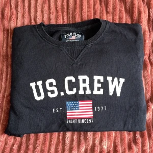 Svart US.CREW tröja Polo Club - Svart tröja från Polo Club med US.CREW-tryck och amerikansk flagga på bröstet. Rund halsringning, ribbade muddar och klassisk collegekänsla. Materialet är mjuk bomull, perfekt för en avslappnad och sportig stil.