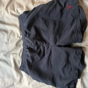  Gant Badbyxor - Svarta shorts från Gant med elastisk midja och diskret röd logga broderad framtill. Klassisk och enkel design, perfekta för sommardagar eller träning. Lätta och bekväma att bära, passar till det mesta.