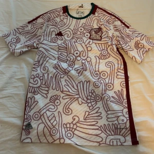 Mexiko fotbollströja Adidas mönstrad - Snygg Mexiko fotbollströja från Adidas med unikt vinrött mönster på vit bas. Tröjan har korta ärmar, grönt kantband vid halsen och broderad Mexiko-logga på bröstet. Perfekt för dig som vill sticka ut på match eller träning. Använd några gånger och är o bra skick.