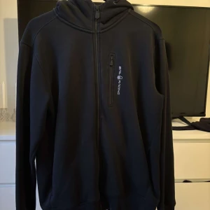 Svart hoodiejacka Sail Racing XL - Svart hoodiejacka från Sail Racing i storlek XL. Jackan har hel dragkedja framtill, huva med snörning och en bröstficka med dragkedja. Snygg logga på bröstet och en clean look som passar till det mesta. Perfekt för dig som gillar sportig och stilren stil.