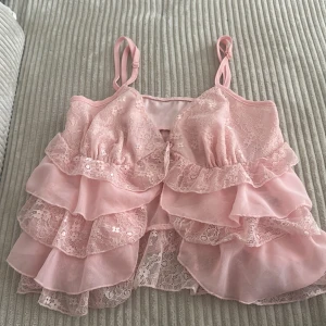 Rosa volangig linnetopp med spets - Supersöt rosa linnetopp med flera lager volanger och spetsdetaljer. Axelbandsmodell med romantisk känsla, perfekt för sommardagar. Tunn och luftig, passar till både jeans och kjol.