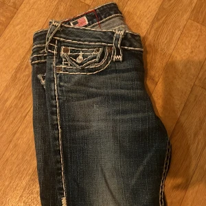 True Religion jeans med kontrastsömmar - Säljer ett par blå jeans från True Religion med tydliga vita kontrastsömmar och stora fickor med dekorativa knappar. Klassisk femficksmodell och ikonisk logga bak. Snygga detaljer som ger en unik look.