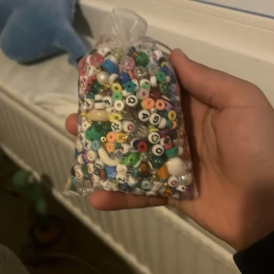Påse med färgglada pärlor - En påse fylld med blandade färgglada pärlor i olika former och storlekar, perfekta för att göra egna smycken som armband, halsband eller ringar. Pärlorna har bland annat bokstäver, ögon och olika mönster.
