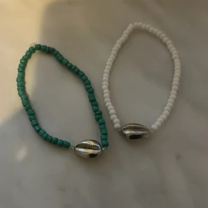 Två armband med pärlor och snäckdetalj - Två armband med små pärlor, ett i grönt och ett i vitt. Båda har en silverfärgad snäckformad detalj. Perfekta som accessoar för en somrig look.
