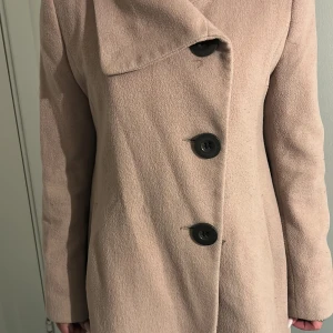 Beige kappa från Isolde - Säljer en klassisk beige kappa från Isolde i storlek 38. Jackan har stora svarta knappar framtill, bred krage och ett bälte baktill för snygg passform. Perfekt för höst och vår, stilren och enkel att matcha.