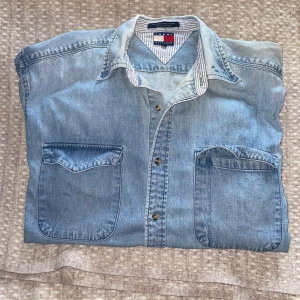 Ljusblå jeansskjorta från Tommy Hilfiger - Säljer en klassisk ljusblå jeansskjorta från Tommy Hilfiger med två stora bröstfickor och knappar framtill. Skjortan har lång ärm och en snygg krage med randigt innerfoder. Perfekt för en avslappnad och stilren look.