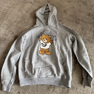 Grå hoodie med björntryck från Moschino - Säljer en grå hoodie från Moschino med ett stort tryck av en söt björn och texten 'TOY' på framsidan. Tröjan har huva och ribbade muddar vid ärmslut och nederkant. Perfekt för en chill och trendig look.