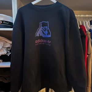 Svart sweatshirt från Adidas Adventure - Säljer en svart sweatshirt från Adidas Adventure med tryck av en lila björn och text på bröstet. Tröjan har rund halsringning och långa ärmar. Perfekt för en avslappnad stil.