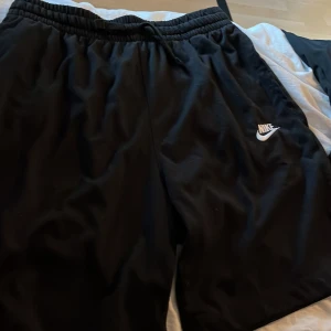 Svarta träningsshorts från Nike - Svarta shorts från Nike med elastisk midja och vit logga på vänster ben. Perfekta för träning eller sportiga aktiviteter. Lätta och bekväma med normal passform.