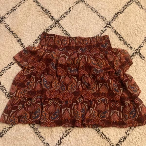 Mönstrad volangkjol från Lindex - Säljer en röd mönstrad volangkjol från Lindex med paisleyinspirerat tryck i orange, blått och rosa. Kjolen har resår i midjan och flera lager volanger för en extra flowy känsla. Strl 158\164 men skulle säga att den passar många fler strl pågrund av stretchigt material❣️ t ex passar den mig som vanligt vid har xs-m