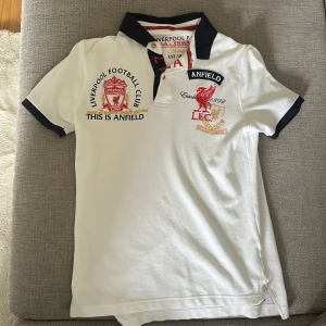 Vit piké t-shirt från Liverpool FC - Vit piké t-shirt med broderade Liverpool FC-loggor och textdetaljer på bröstet. Svart krage och ärmslut, samt knappar vid halsen. Perfekt för fotbollsfans som vill visa sitt stöd för laget.