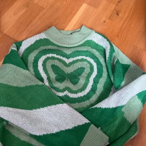 Grön och vit stickad tröja med fjärilsmotiv - Säljer en cool stickad tröja i grönt och vitt med breda ränder och ett stort fjärilsmotiv på bröstet. Tröjan har rund hals och långa ärmar. Perfekt för dig som vill sticka ut lite extra!