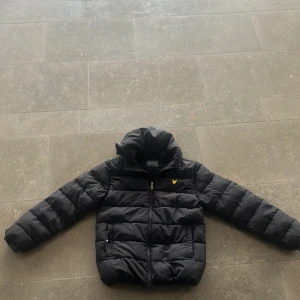 Svart pufferjacka från Lyle & Scott - Säljer en svart pufferjacka från Lyle & Scott med huva och dragkedja. Jackan har det klassiska gula logobroderiet på bröstet och två fickor framtill. Perfekt för kyliga dagar.