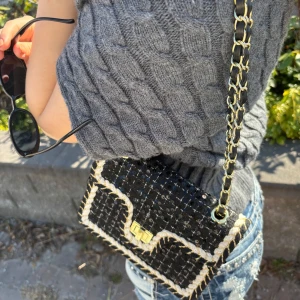 Svart axelväska med kedjerem och guldiga detaljer - Snygg svart axelväska med rutigt mönster och guldiga detaljer. Väskan har en kedjerem i guld och svart, samt ett dekorativt lås framtill. Perfekt storlek för det viktigaste och ger en trendig touch till outfiten.