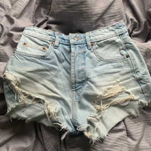Ljusblå slitna jeansshorts från Zara - Säljer ett par ljusblå jeansshorts från Zara med hög midja och råa, slitna kanter. Shortsen har klassiska fem fickor, knappgylf och fransiga detaljer framtill för en cool, avslappnad look.