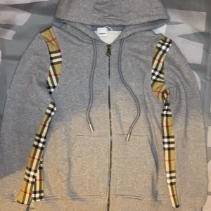 Grå hoodie med rutiga detaljer - Grå hoodie med dragkedja och huva, dekorerad med klassiska beige, svarta och röda rutiga band längs ärmarna. Två fickor framtill och justerbar dragsko i huvan. Perfekt för en avslappnad stil.