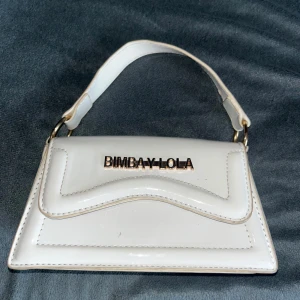 Vit handväska från BIMBA Y LOLA - Säljer en stilren vit handväska från BIMBA Y LOLA med guldfärgade detaljer och bredt handtag. Väskan har en unik form och logotypen i metall framtill. Perfekt accessoar för att lyfta din outfit.