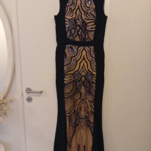 Svart och guldig långklänning med paljetter - Säljer en elegant långklänning i svart med guldiga paljettmönster framtill. Klänningen är ärmlös och har en figurnära passform med snygga detaljer som ger en lyxig känsla. Perfekt för bal eller festliga tillfällen.