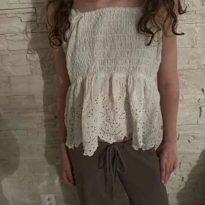 Vit topp med volang och broderade detaljer - Säljer en söt vit topp med smock upptill och volang nertill. Toppen har fina broderade hålmönster längs volangen. Perfekt för varma dagar och ger en romantisk känsla. Pris kan diskuteras.