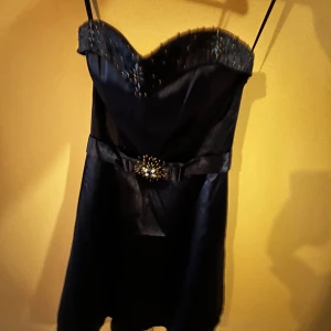  axelbandslös klänning med strass och rosettdetalj - Säljer en blå axelbandslös klänning med smala axelband, strassdetaljer upptill och snygg rosett med dekoration i midjan. Klänningen har snörning i ryggen och är perfekt för festliga tillfällen.