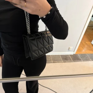 Svart quiltad axelväska med kedja - Säljer en svart axelväska med quiltat mönster och silverfärgad kedjerem. Väskan har en stilren, rektangulär form och stängs med magnetknappar. Perfekt för att bära det viktigaste och ger en trendig touch till din outfit. Den är väldigt rymlig och får plats med allt du behöver ha med dig!!🖤