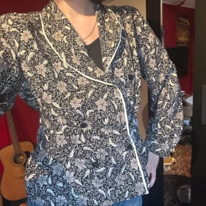 Svartvit blommig kavaj med kontrastkant - Säljer en svartvit kavaj med blommigt mönster och vita kontrastkanter. Dubbelknäppt framtill och med långa ärmar. Perfekt för dig som vill sticka ut med en unik och stilren look.