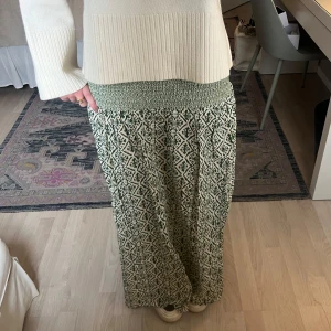 Grönmönstrad långkjol - Säljer en långkjol med grönt och vitt grafiskt mönster och bred smock i midjan. Kjolen är luftig och har en avslappnad passform, perfekt för varmare dagar. Mönstret ger en bohemisk känsla och midjan är elastisk för extra komfort. Aldrig använd!